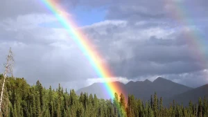 A vibrant rainbow stretches across a peaceful mountain forest beneath dramatic clouds, symbolizing enlightenment, Truth, and the path to human completion through Ma-eum Su-Ryun Meditation.
